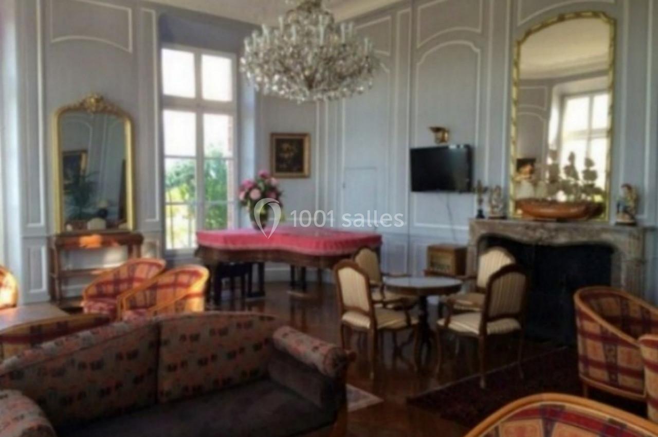 Salon élégant avec moulures, lustre, cheminée, miroirs, table recouverte d’un tissu rouge et mobilier classique.