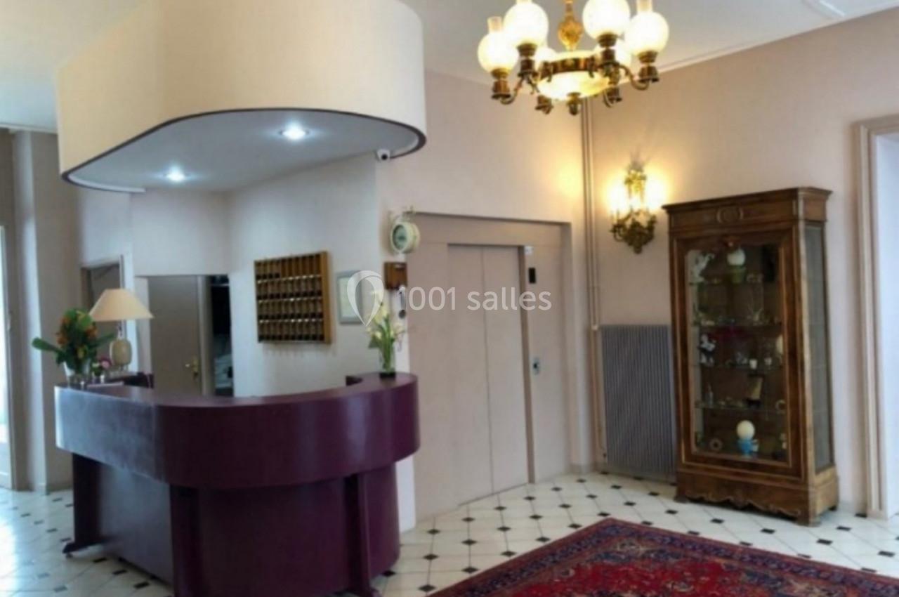 Accueil d'un hôtel avec comptoir violet, lustre ancien, vitrine en bois et tapis rouge sur un sol carrelé.