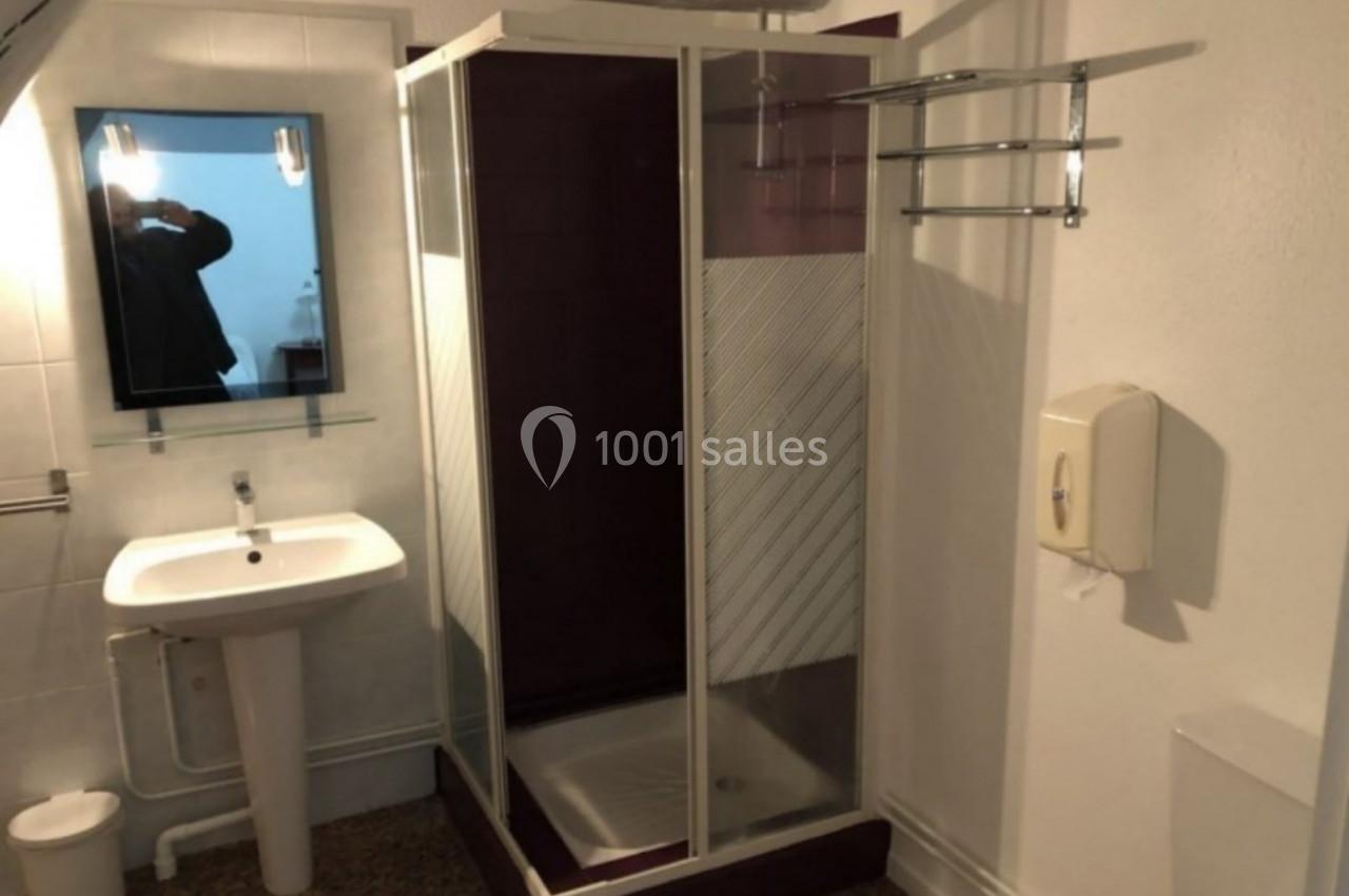 Salle de bain avec douche d'angle, lavabo avec miroir, porte-serviettes mural et distributeur de savon.