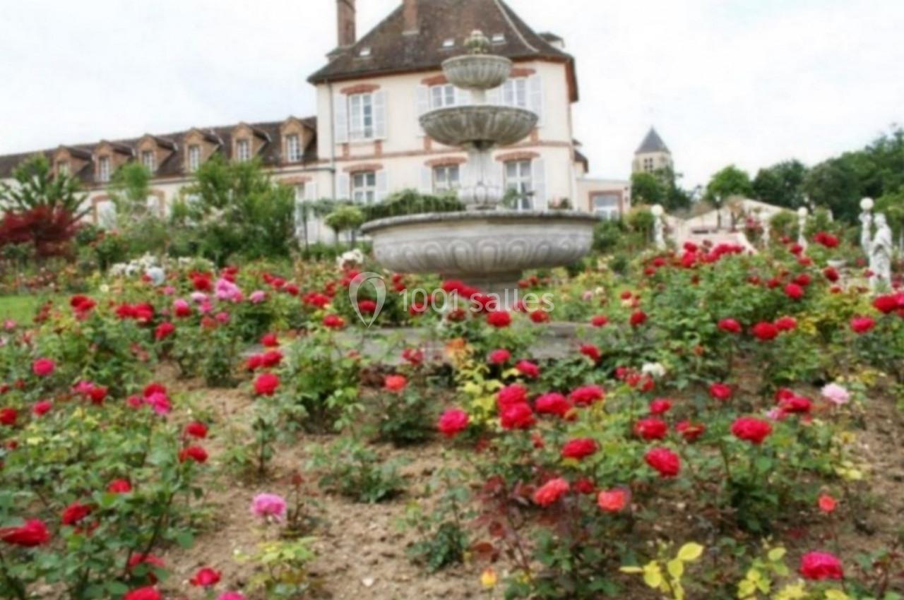 Jardin fleuri avec des rosiers colorés entourant une fontaine en pierre devant une grande maison ancienne.