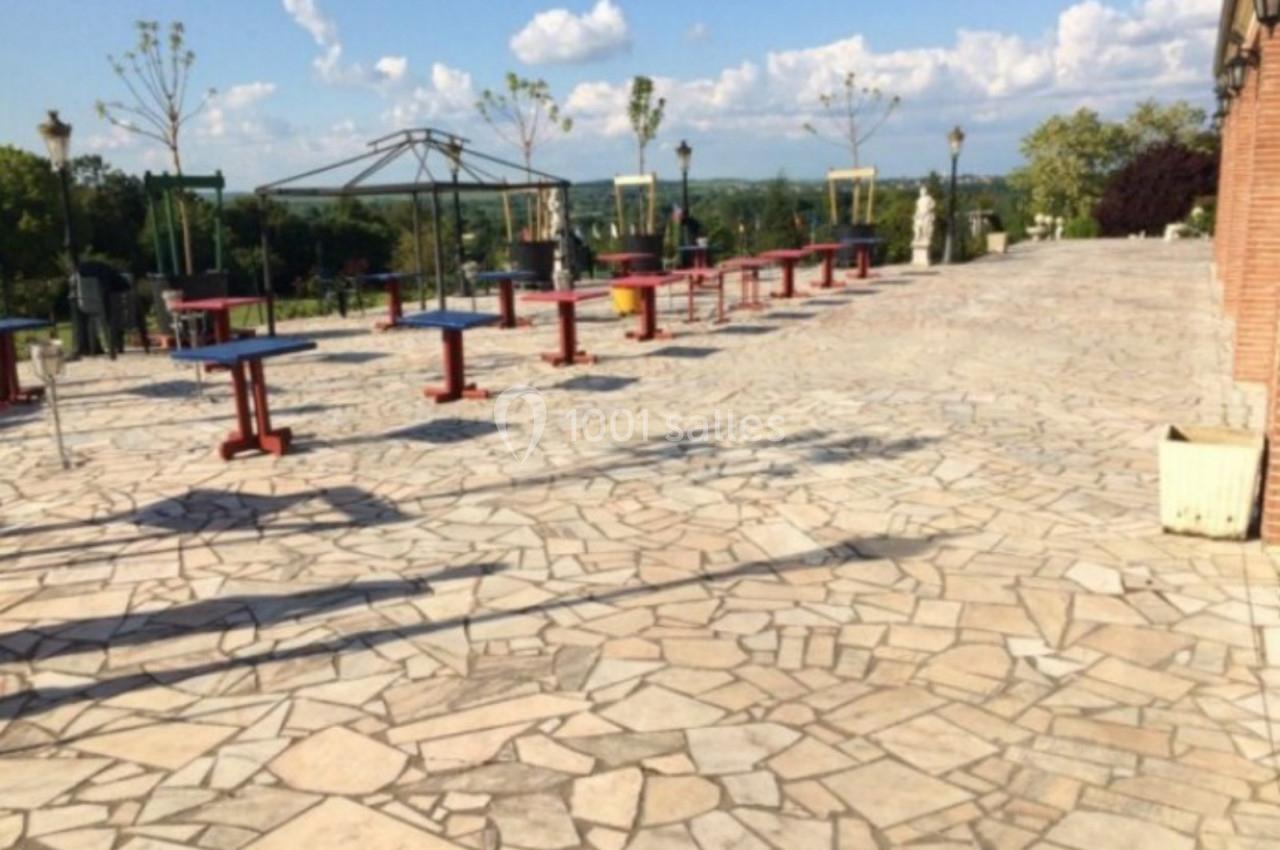 Terrasse pavée avec des tables colorées espacées, entourée de verdure et offrant une vue dégagée sur l'horizon.