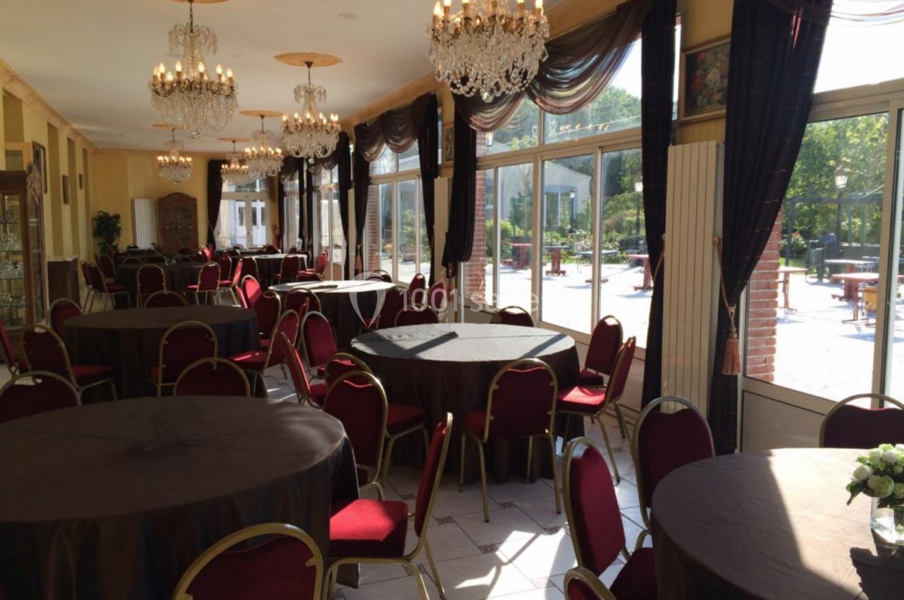 Salle de réception lumineuse avec tables rondes nappées de noir, chaises rouges et lustres en cristal.
