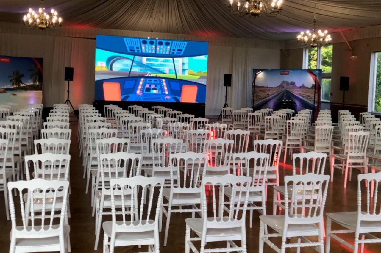 Salle de conférence avec des rangées de chaises blanches face à un écran géant affichant une scène de cockpit.