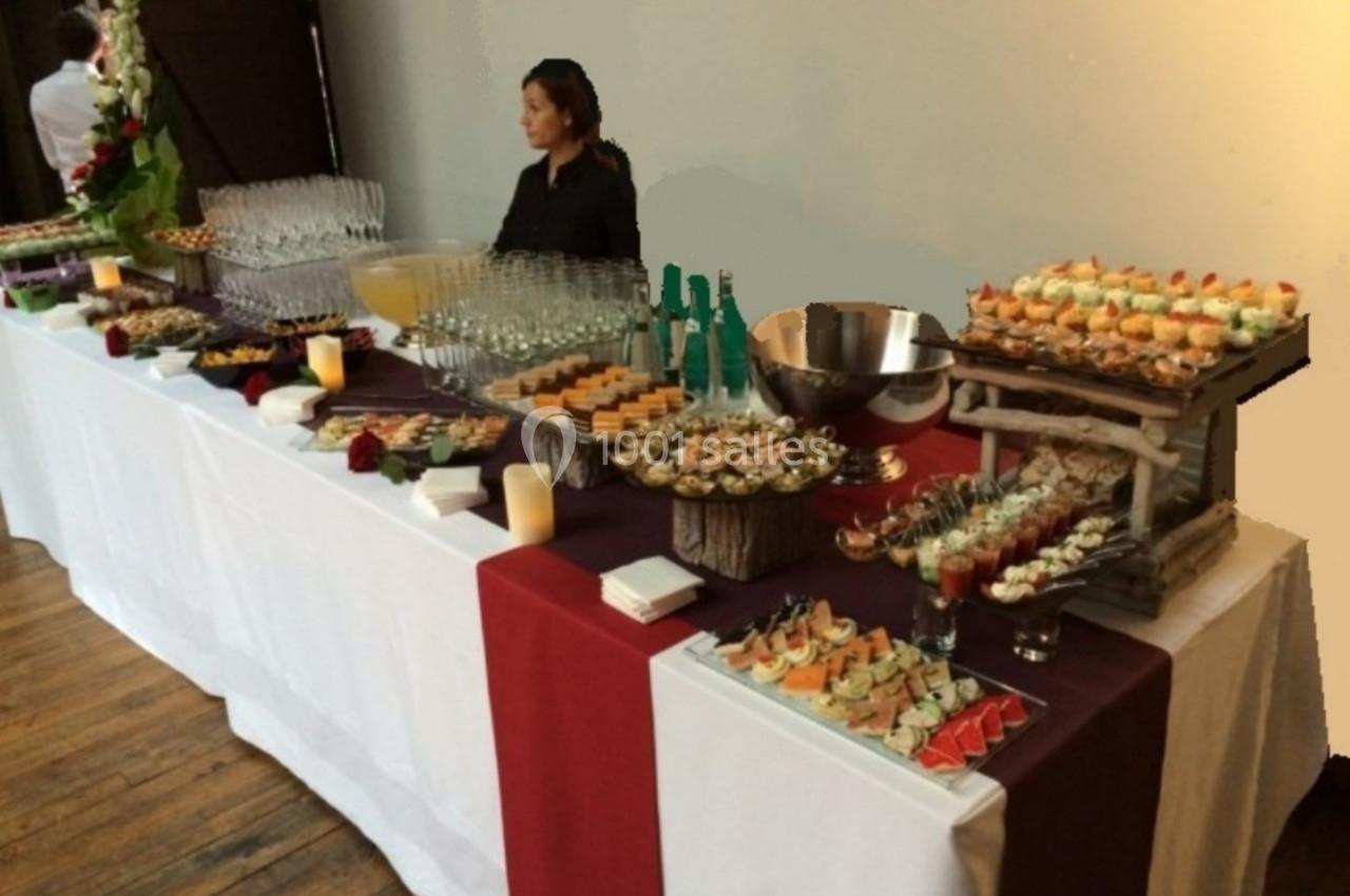 Table de buffet avec verrines, amuse-bouches variés, boissons et décorations florales, une personne en arrière-plan.