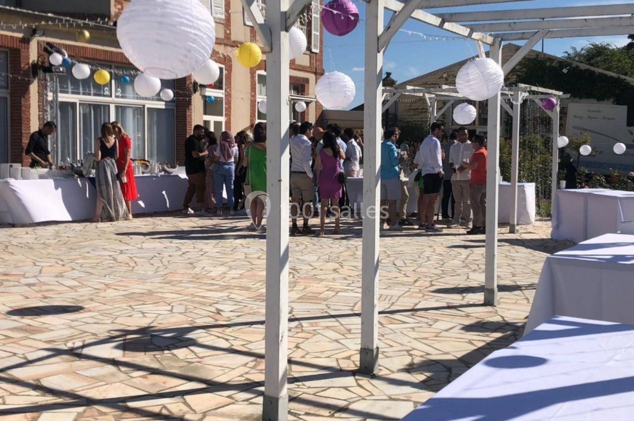 Groupe de personnes rassemblées sur une terrasse ensoleillée décorée de lanternes blanches et de tables dressées.