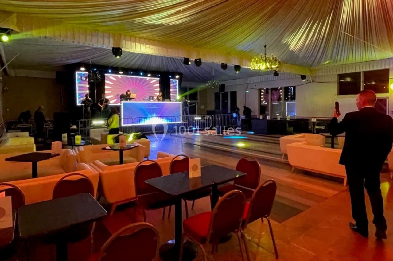 Salle de réception avec tables, fauteuils et scène éclairée, préparée pour un événement avec jeux de lumières.