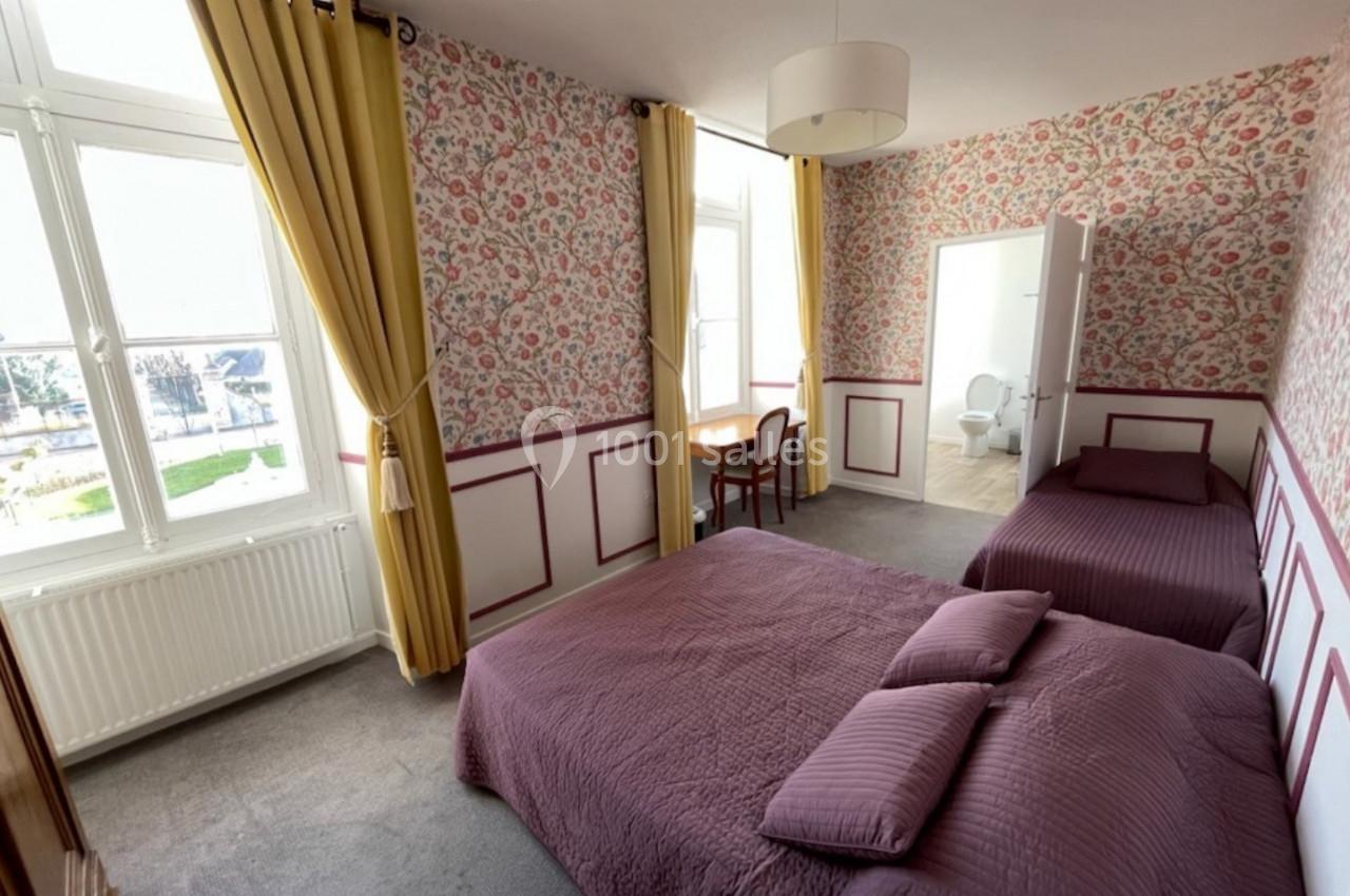 Chambre lumineuse avec deux lits, murs à motifs floraux, rideaux jaunes et vue sur l'extérieur par de grandes fenêtres.