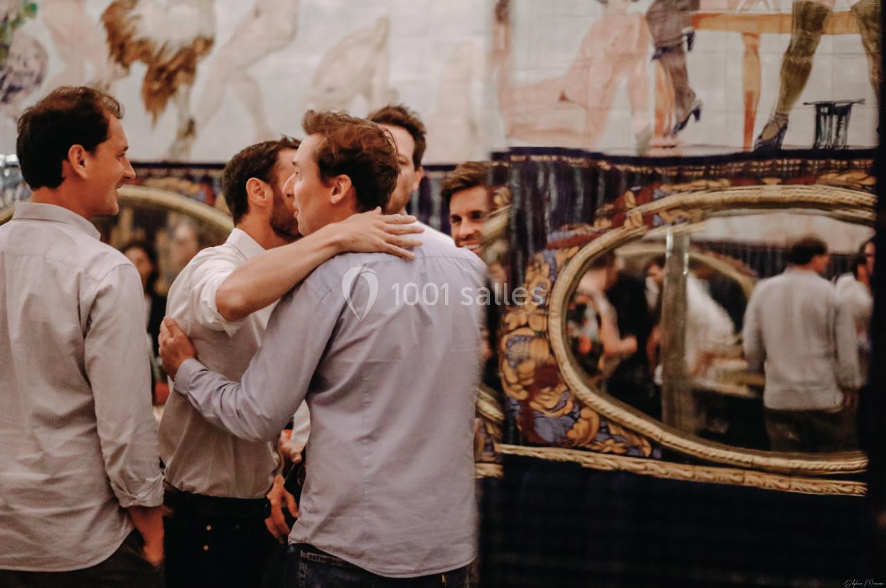 Un groupe d'hommes discutant et s'embrassant dans un intérieur décoré avec des miroirs et des motifs artistiques.