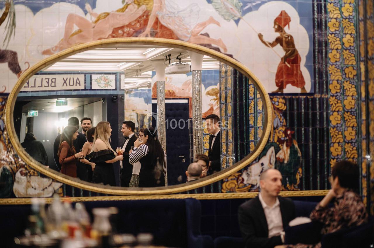 Un groupe de personnes discutant dans un intérieur décoré de fresques colorées, reflété dans un grand miroir ovale.