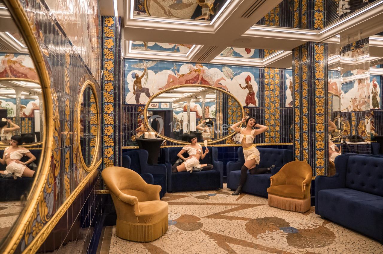 Deux femmes posent dans un salon richement décoré avec des miroirs, des fresques colorées et des fauteuils élégants.