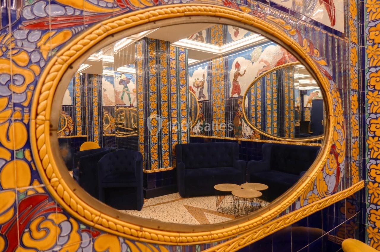 Miroir ovale reflétant un salon avec canapés bleus, tables basses et murs ornés de carreaux colorés.