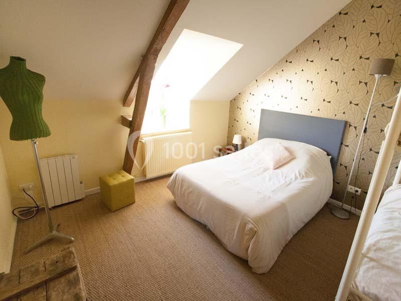 Chambre mansardée avec lit double, mur tapissé, mannequin de couture et lumière naturelle provenant d'une fenêtre.