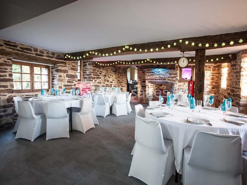 Salle en pierre avec tables rondes dressées de nappes blanches, chaises blanches et décorations lumineuses suspendues.