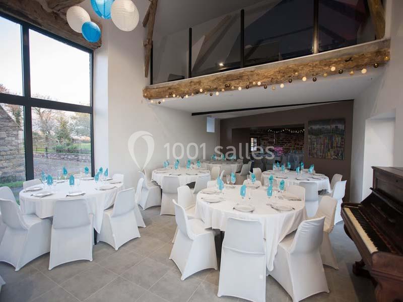 Salle de réception lumineuse avec tables rondes dressées, chaises blanches, et mezzanine en bois visible en arrière-plan.