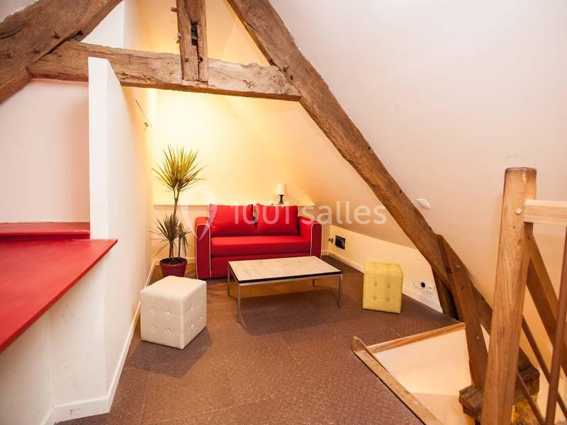 Salon mansardé avec poutres apparentes, canapé rouge, table basse, poufs et éclairage doux.