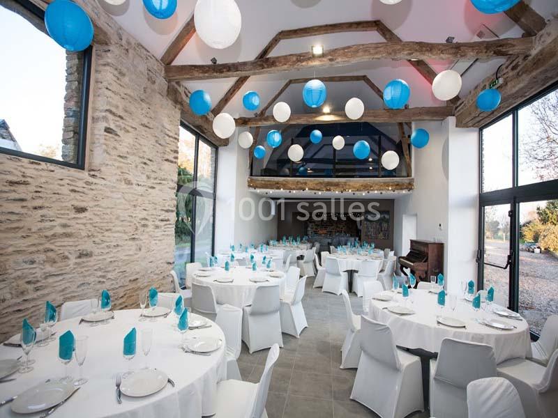 Salle de réception lumineuse avec tables rondes dressées, décorée de lanternes bleues et blanches suspendues au plafond.