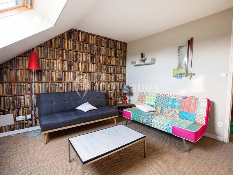 Salon mansardé avec canapé gris, canapé coloré, table basse en bois et mur tapissé d’un motif bibliothèque.