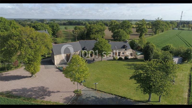 Vue aérienne d'une maison en pierre avec toit en ardoise, entourée d'un grand jardin et de champs verdoyants.