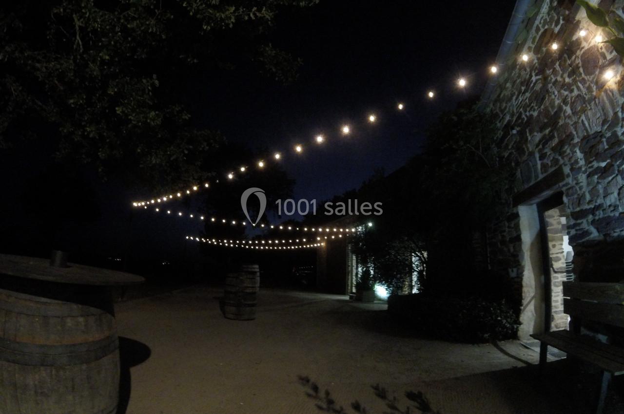 Guirlandes lumineuses suspendues au-dessus d'une cour extérieure sombre, bordée d'un bâtiment en pierre et de tonneaux.