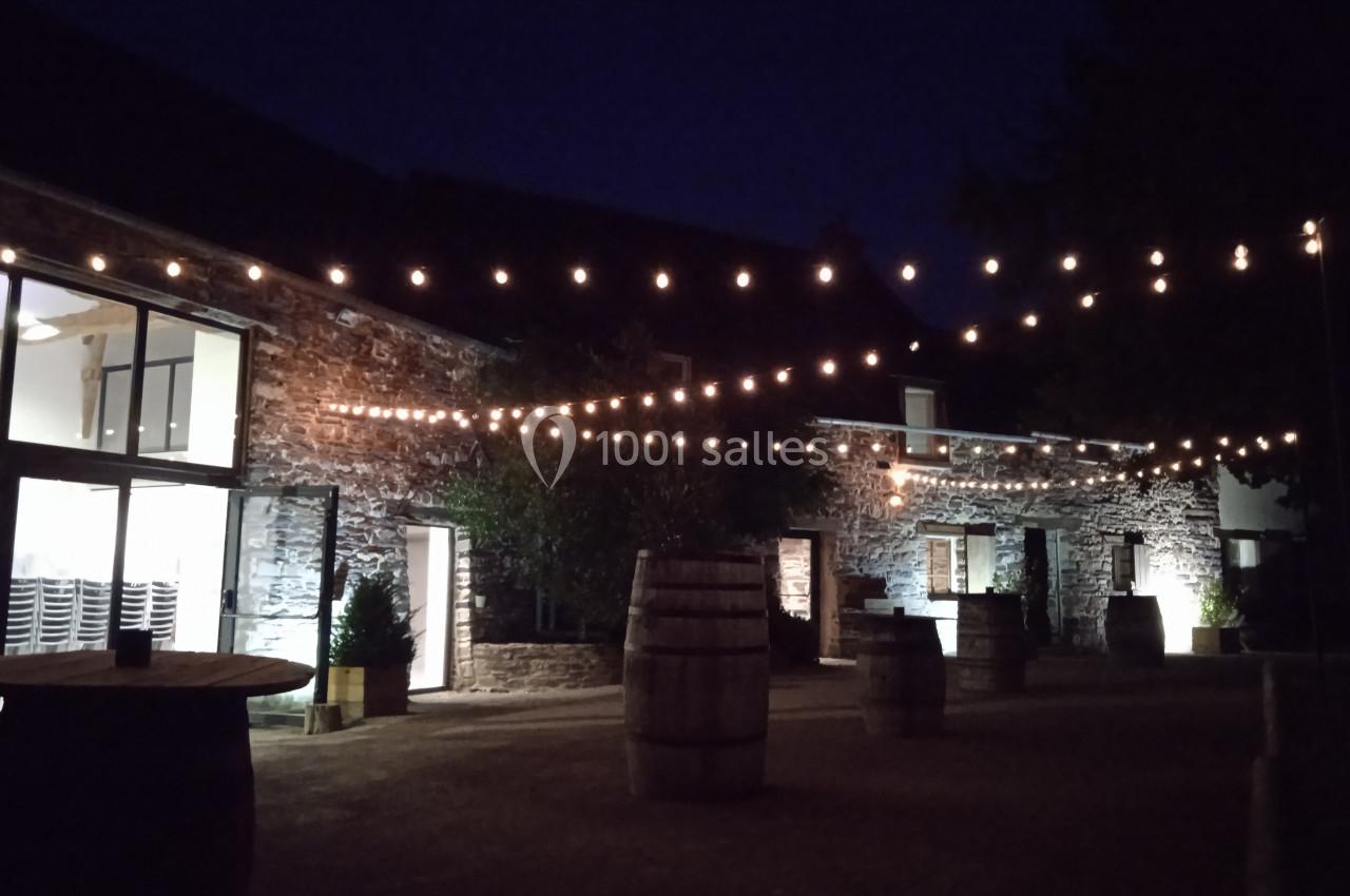 Cour extérieure en soirée avec guirlandes lumineuses suspendues, tonneaux en bois et bâtiments en pierre éclairés.