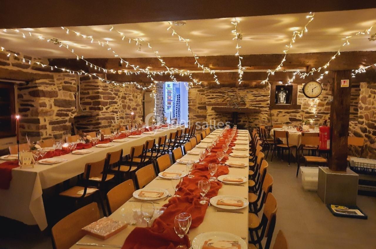Salle décorée avec des guirlandes lumineuses, tables alignées et dressées pour un repas dans un cadre en pierre.