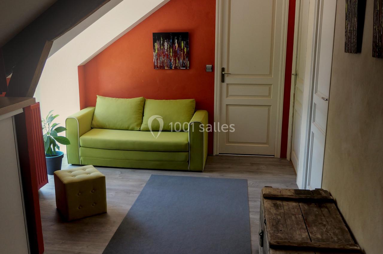 Couloir mansardé avec un mur orange, un canapé vert, des coffres en bois et une lumière naturelle provenant d'une fenêtre.