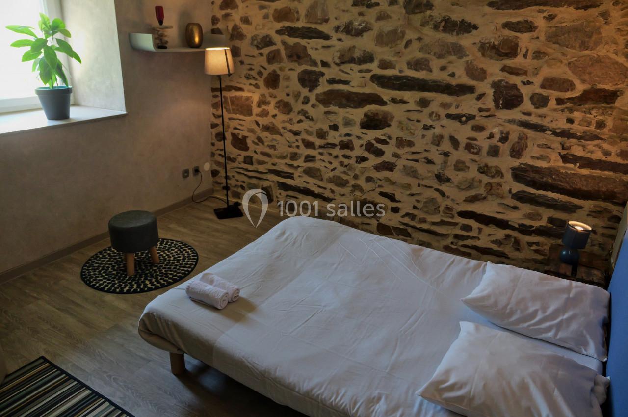 Lit bas avec draps blancs dans une chambre à mur en pierre, éclairée par une fenêtre et une lampe sur pied.