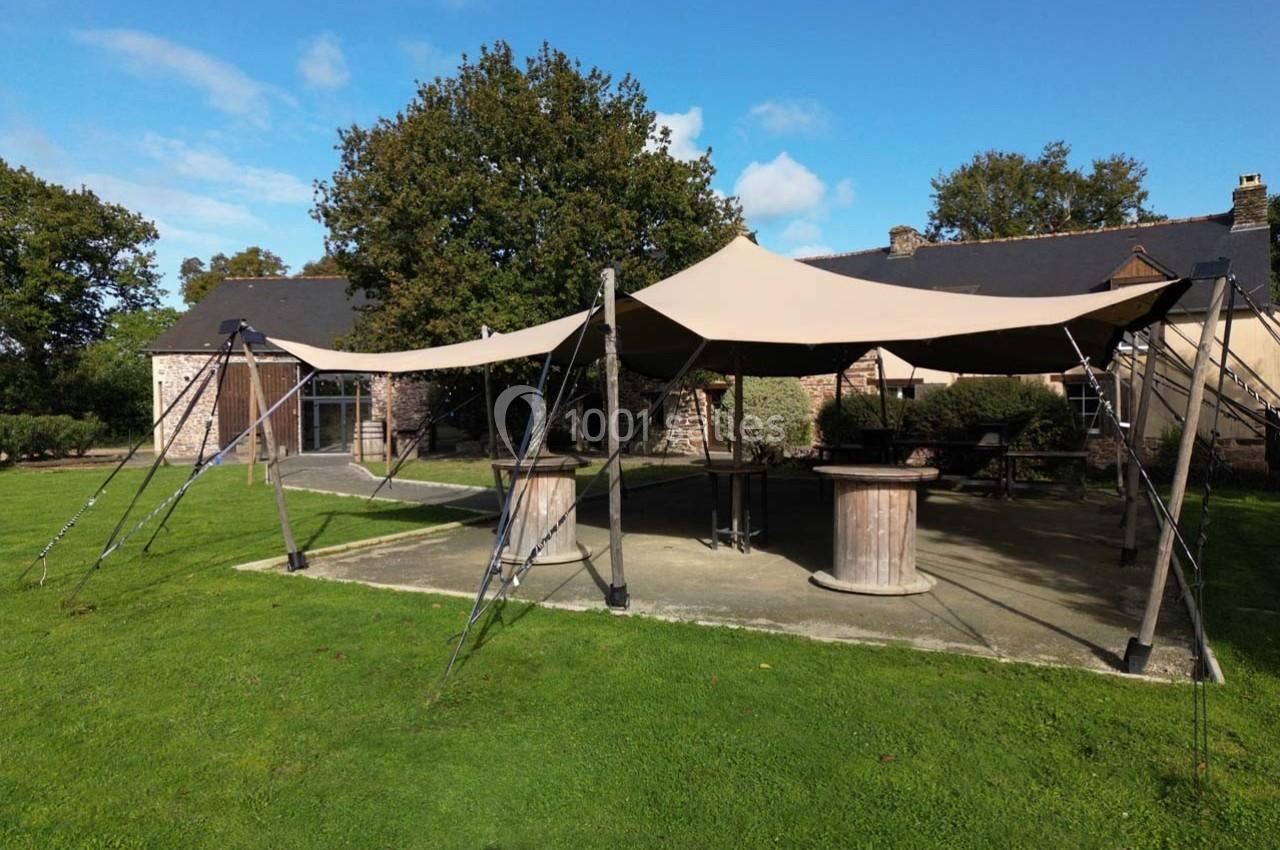 Tente beige montée sur une terrasse en pierre, entourée de verdure et d'arbres, devant un bâtiment en pierre.