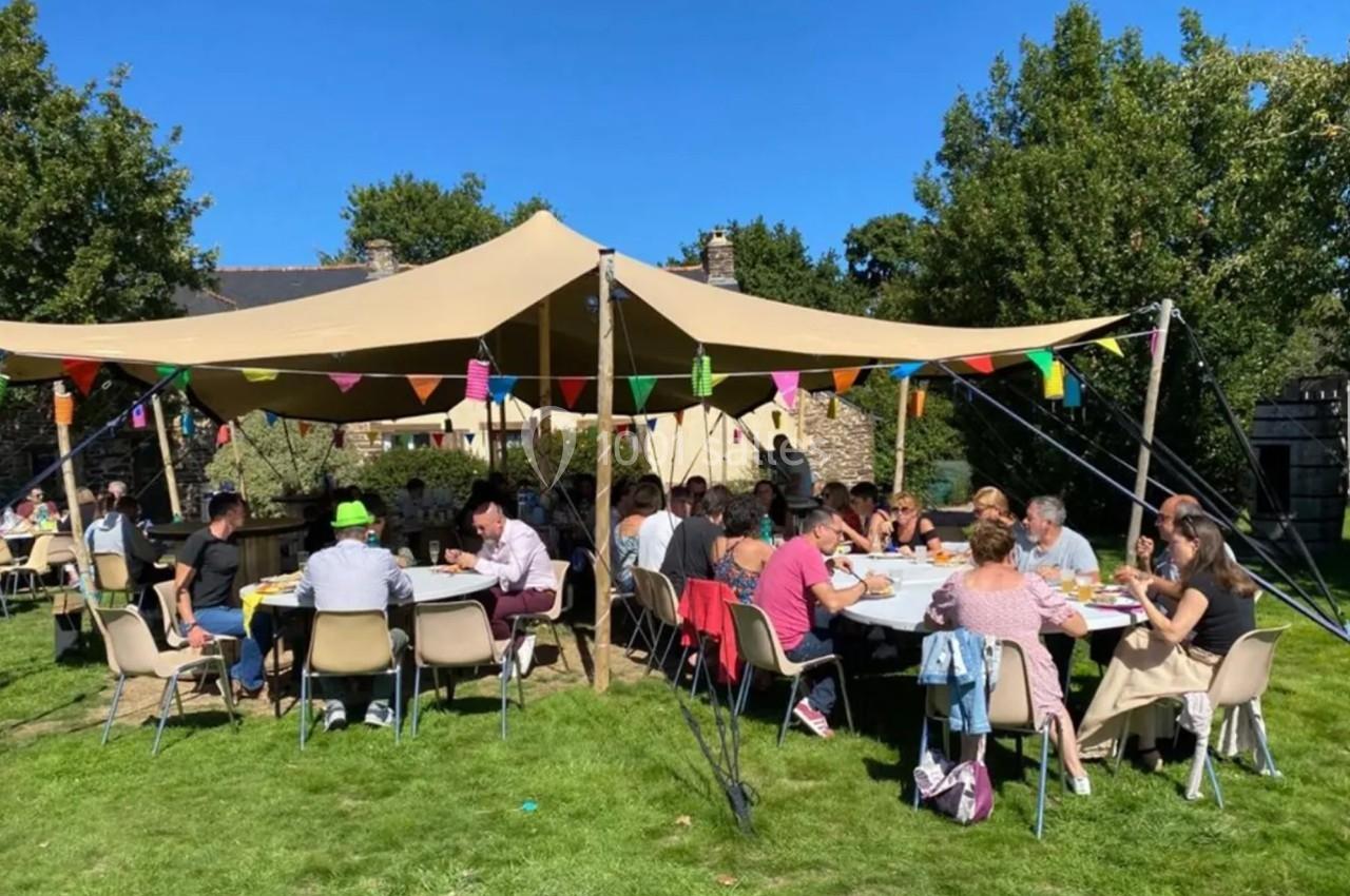 Des personnes assises à des tables rondes sous une grande tente beige, dans un jardin ensoleillé avec des guirlandes…