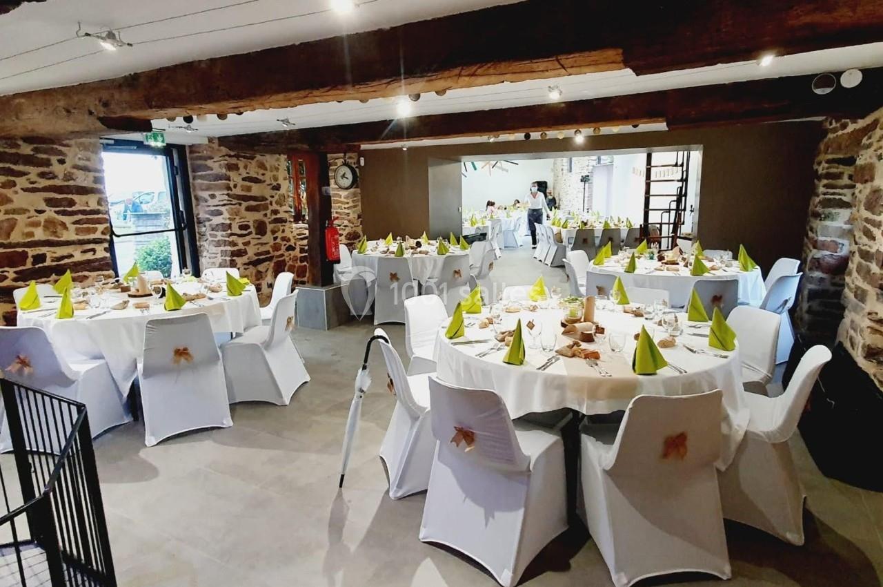 Salle de réception décorée avec des tables rondes, nappes blanches, serviettes vertes et murs en pierre apparente.