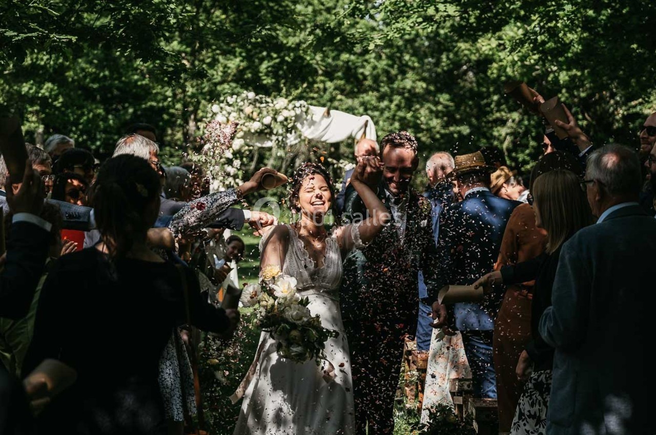 Un couple souriant marche sous une pluie de confettis lors d'une cérémonie en plein air entourée d'invités.