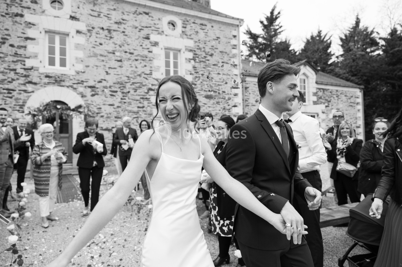 Un couple souriant sort d'un bâtiment en pierre, entouré d'invités applaudissant lors d'une cérémonie.