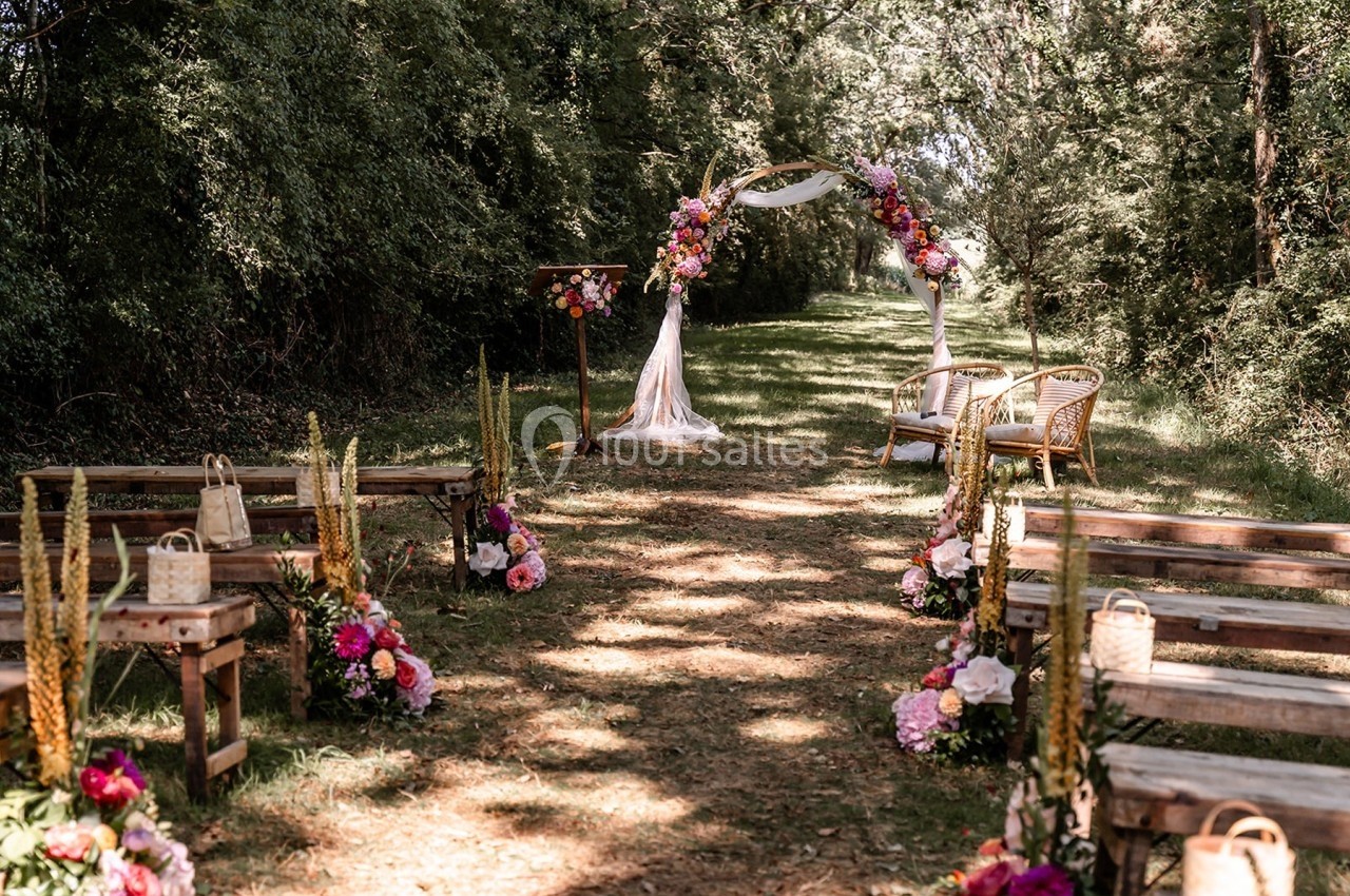Allée décorée de fleurs menant à une arche nuptiale en extérieur, entourée d'arbres et de bancs en bois.