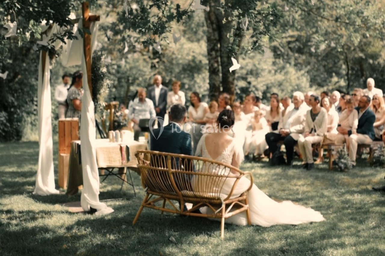 Un couple assis sur un banc en osier lors d'une cérémonie de mariage en plein air, entouré d'invités.