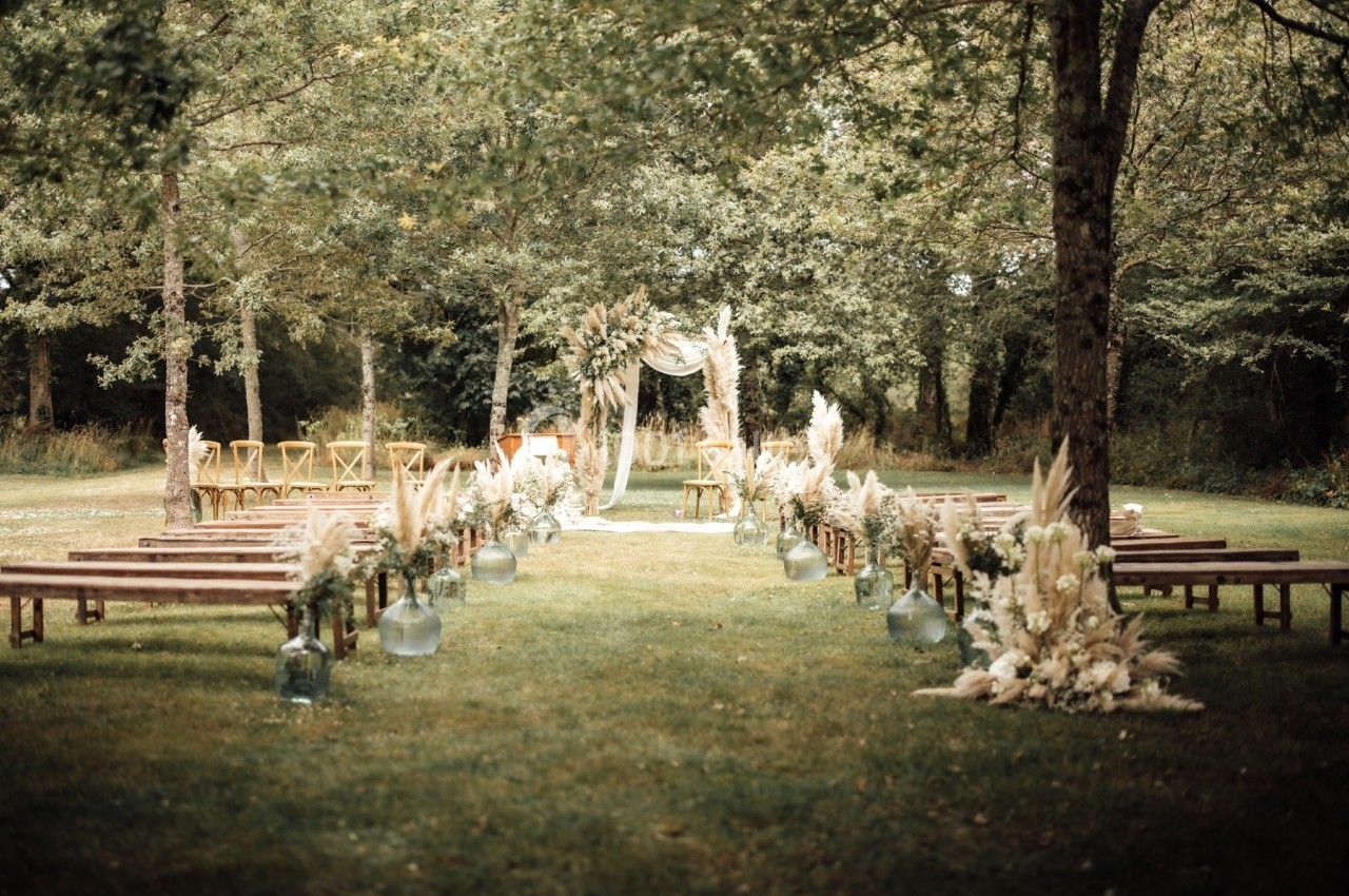 Allée décorée de fleurs et bancs en bois menant à une arche pour une cérémonie en plein air dans un cadre boisé.