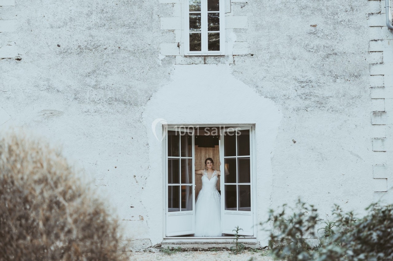 Une femme en robe blanche se tient dans l'encadrement d'une porte-fenêtre ouverte d'une maison en pierre.