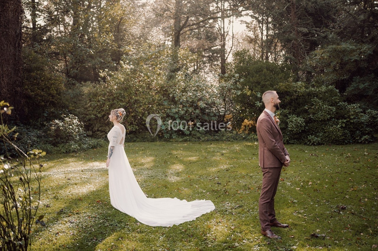 Une mariée en robe blanche et un marié en costume brun se tiennent dos à dos dans un jardin verdoyant.