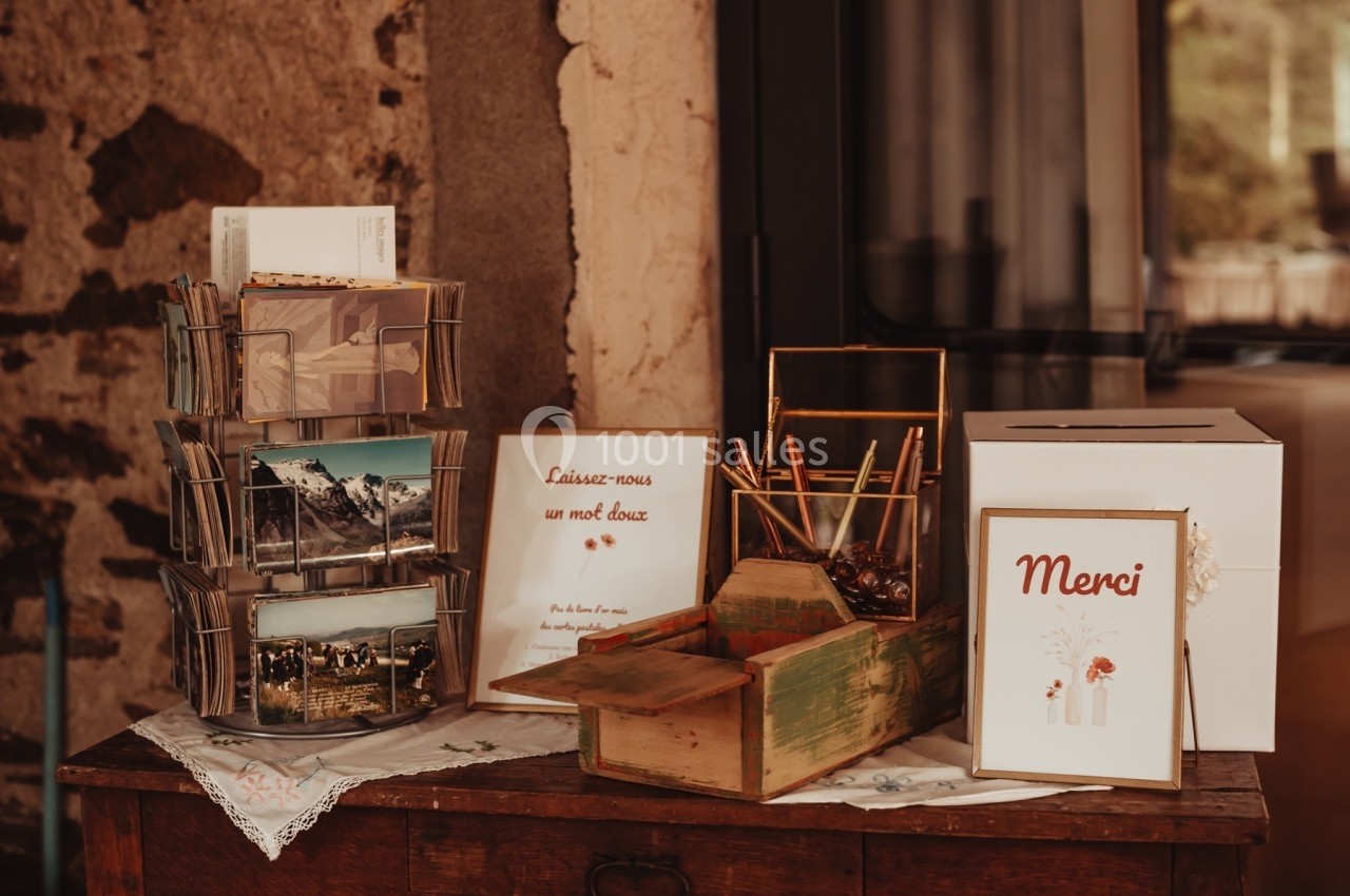 Table avec présentoir à cartes postales, boîte à messages, stylos et pancarte ’Merci’ dans un décor rustique.