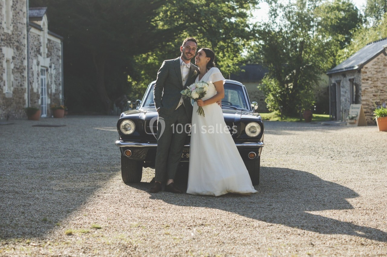 Un couple en tenue de mariage pose devant une voiture ancienne dans une cour ensoleillée bordée de bâtiments en pierre.