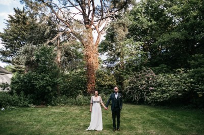 Robe de mariée en dentelle suspendue devant une fenêtre lumineuse avec vue sur un jardin verdoyant.