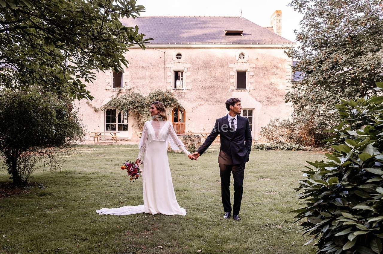 Un couple en tenue de mariage se tient la main dans le jardin d'une maison ancienne en pierre.
