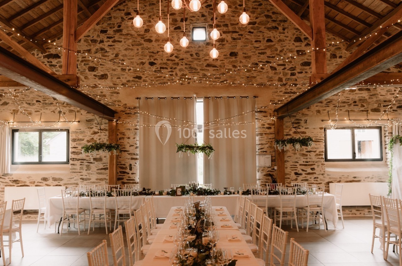 Salle de réception en pierre avec tables décorées, chaises blanches et guirlandes lumineuses suspendues.