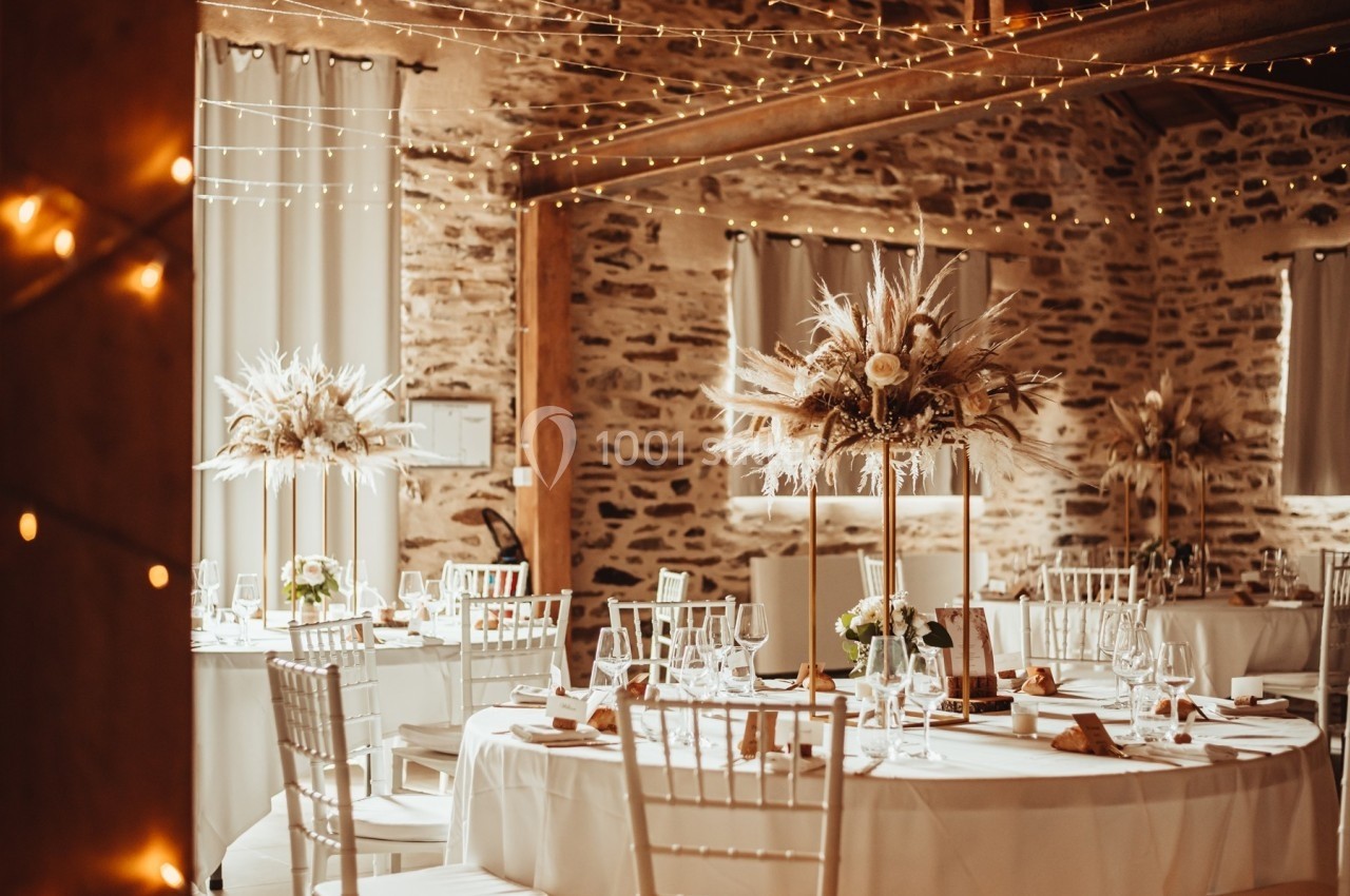 Salle de réception décorée avec des tables rondes, des chaises blanches et des centres de table floraux sous des guirlandes…