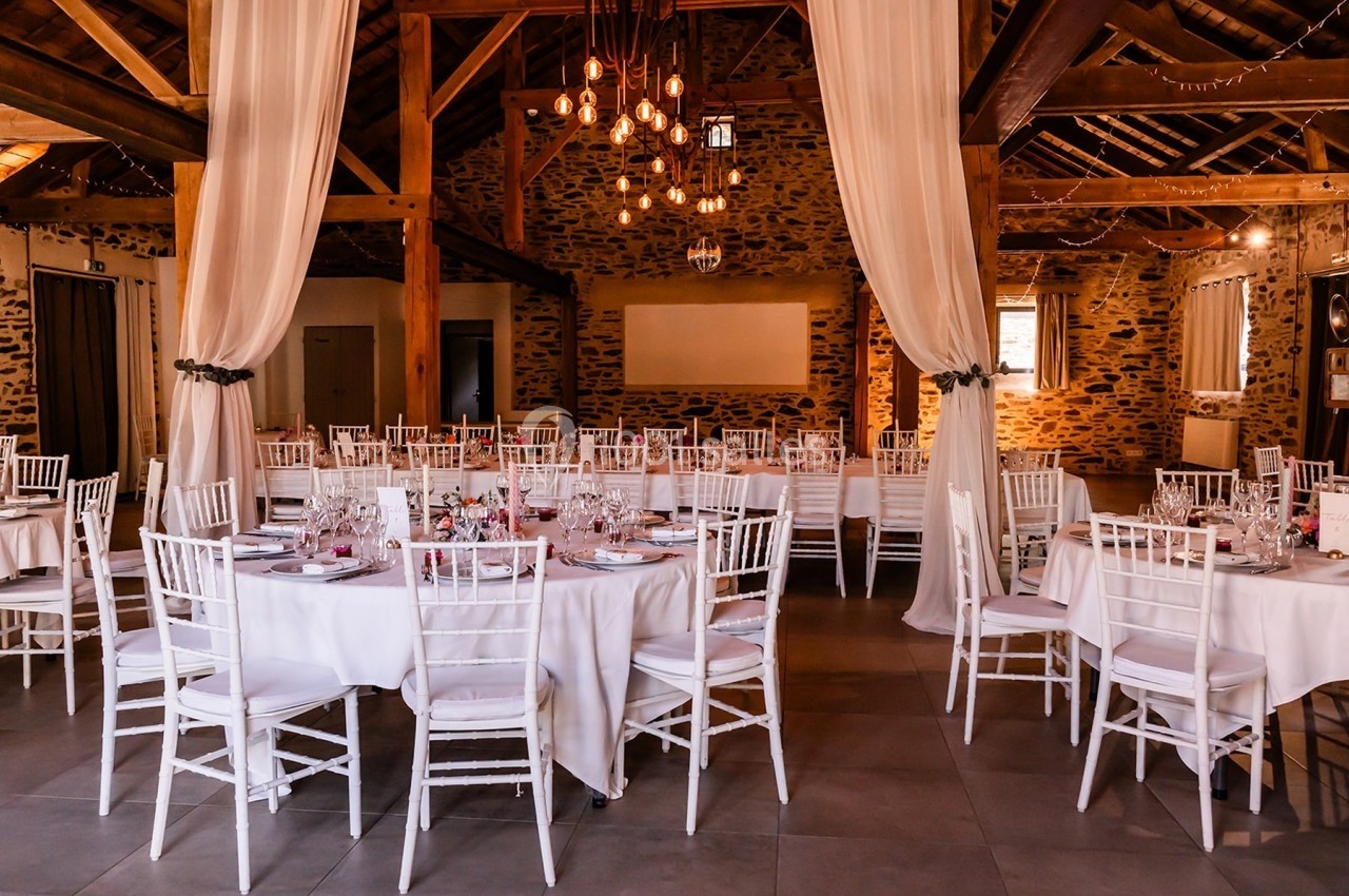 Salle de réception rustique avec murs en pierre, tables rondes dressées et chaises blanches sous un éclairage chaleureux.