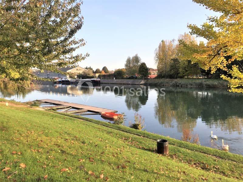 Vue d'une rivière bordée d'arbres en automne, avec un ponton, une barque rouge et des cygnes sur l'eau.