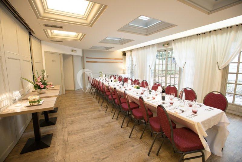 Salle lumineuse avec tables dressées en enfilade, chaises rouges et grandes fenêtres laissant entrer la lumière naturelle.