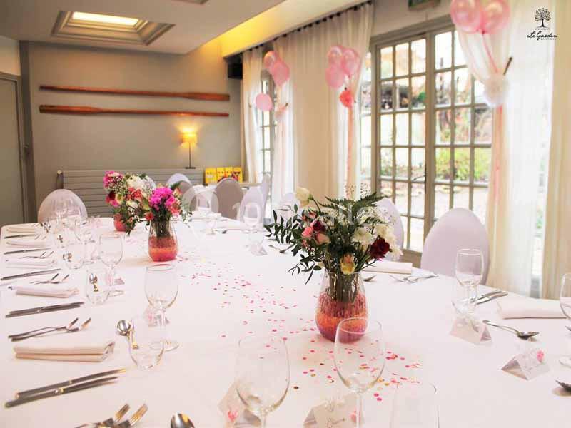 Salle décorée pour un événement, avec une table dressée, des fleurs en centre de table et des ballons roses en arrière-plan.