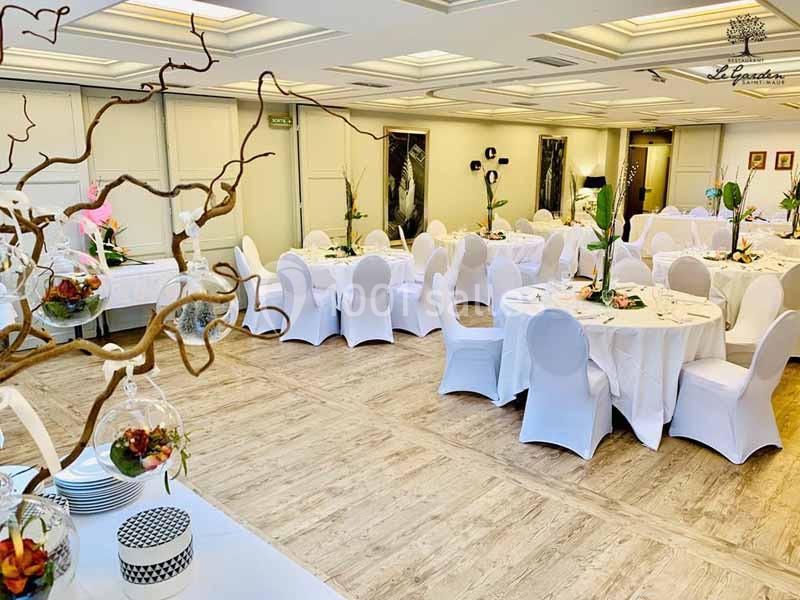 Salle de réception lumineuse avec tables rondes dressées, chaises blanches et décorations florales élégantes.