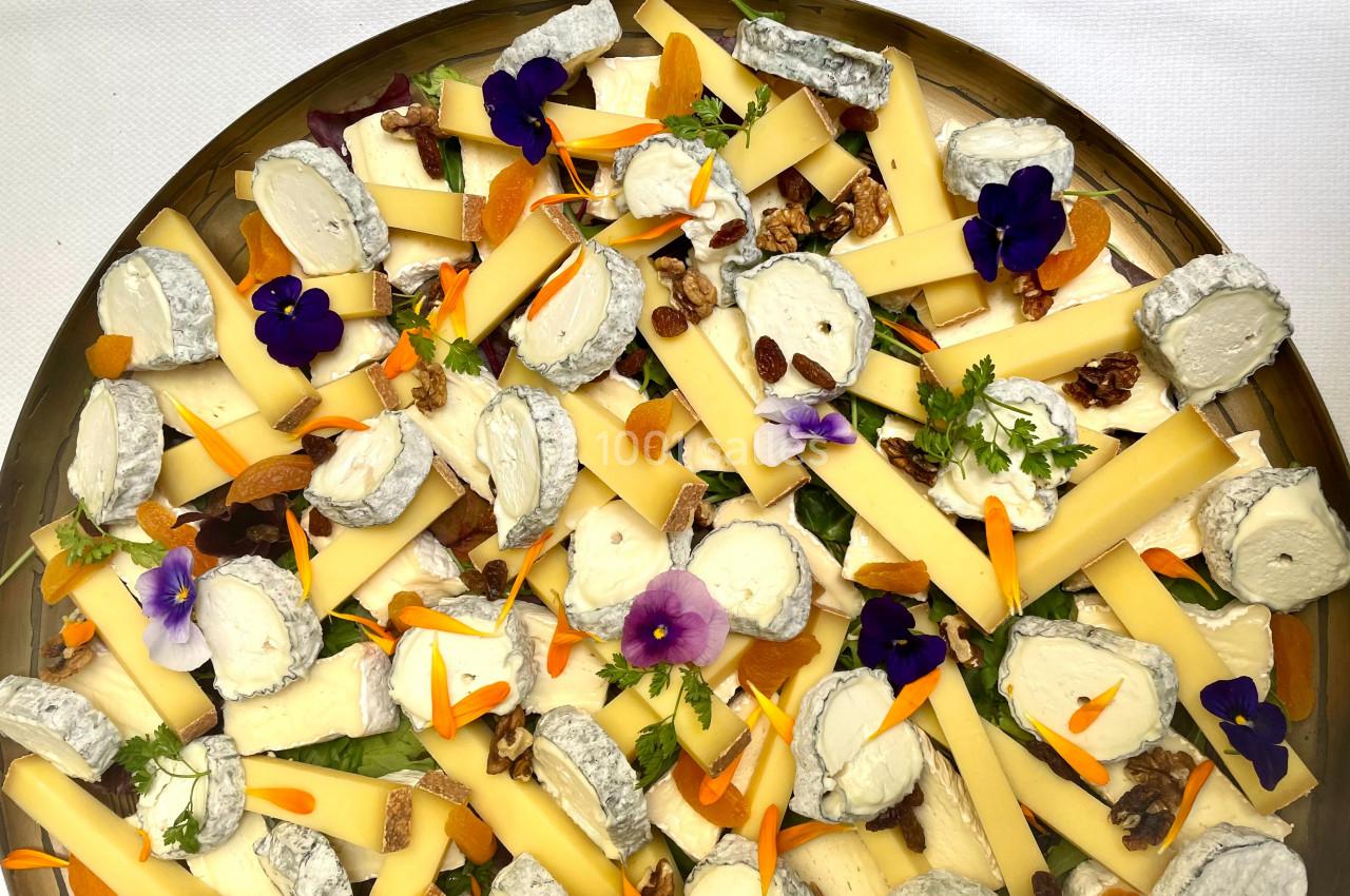 Plateau de fromages variés avec tranches de chèvre, noix, abricots secs et fleurs comestibles sur fond blanc.