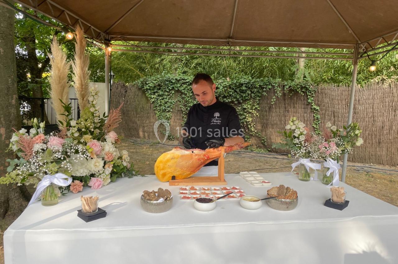Un chef découpe un jambon sur un stand décoré de fleurs, sous une tente en extérieur.